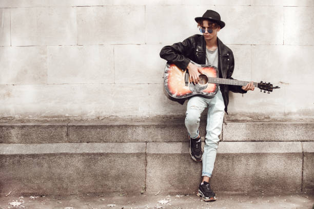 istock Musician. Guy in hat and sunglasses leaning on wall playing guitar thoughtful 1207153272