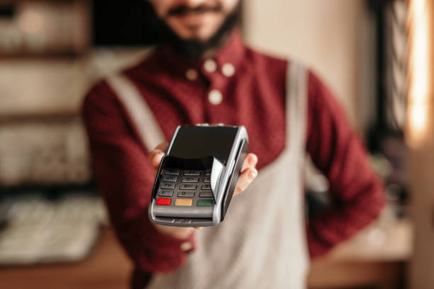 Modern hipster waiter handing over payment terminal in cafe Crop friendly bearded man in uniform holding POS machine for payment with card with empty screen in restaurant point of sale stock pictures, royalty-free photos & images