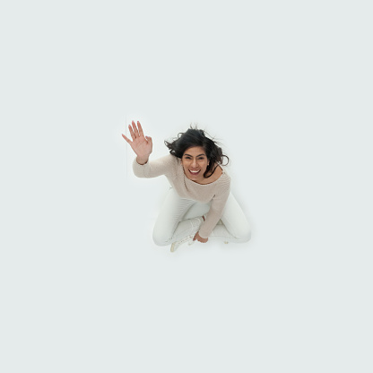 Aerial view of aged 20-29 years old who is beautiful with long hair latin american and hispanic ethnicity young women sitting on floor in front of white background wearing sweater who is happy and greeting and showing hand raised