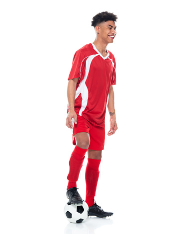 Side view of aged 18-19 years old with curly hair generation z male athlete standing in front of white background wearing soccer uniform who is happy and holding soccer ball and playing soccer - sport and using sports ball