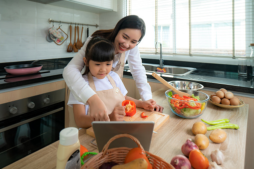 Portrait of beautiful Asian young woman and her daughter cooking salad for lunch using online internet in a digital tablet seach recipe while making food, Family life love relationship, or home fun leisure activity concept