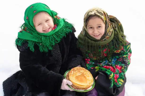 Two charming girls in a Russian folk costume sit on the snow and eat pancakes. The concept of the celebration of the Russian holiday shrovetide