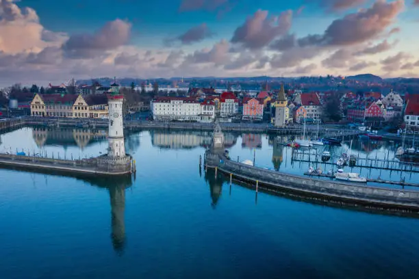 Aerial drone view at sunrise towards the Lindau Island Old Town Cityscape under beautiful twilight skyscape. Lindau Harbor Bodensee - Lake Constance Entrance with the famous old Lighthouse (left) and Bavarian Lion Sculpture from the year 1856 (right). Lindau Waterfront Promenade with Mangenturm, Restaurants and Hotels in the Background. Lindau, Bodensee, Bavaria, Germany.