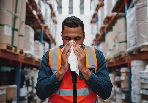 Joven trabajador de almacén africano enfermo soplando la nariz mientras trabaja usando chaleco de seguridad photo