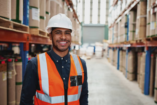 istock Happy male supervisor in warehouse standing in uniform with white hardhat smiling looking at camera - copy space on the right 1207199522