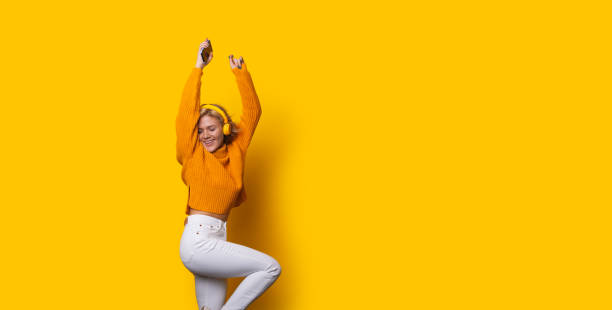 impresionante chica caucásica rubia vestida con un suéter naranja y jeans blancos está bailando mientras posa sobre un fondo amarillo y escucha música - orange white audio fotografías e imágenes de stock