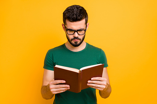 Portrait of focused college student guy hold read textbook learn information for study, project wear spectacles good looking clothes isolated over yellow color background