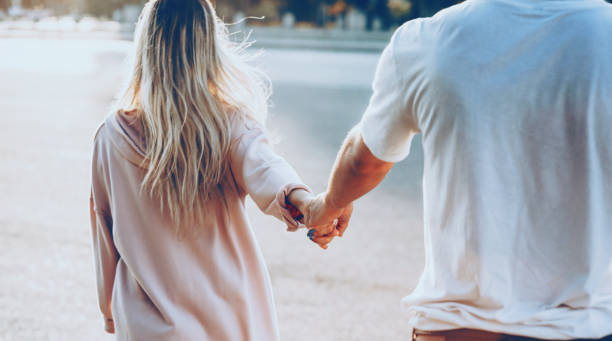 la vista trasera de una joven pareja caucásica vestida de blanco caminando de la mano durante el sol los está calentando - couple human hand holding walking fotografías e imágenes de stock