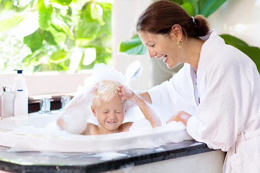 Little child taking bubble bath in beautiful bathroom with big garden view window. Mother washing baby. Kids hygiene. Shampoo, hair treatment and soap foam for children. Mom bathing kid in large tub.