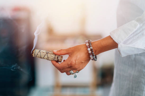 A close up image of a smoldering white sage smudge bundle and healing crystal on a dark wooden table.