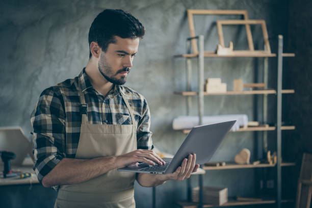 istock Photo of handsome guy holding notebook watching online video masterclass have free self education focused wooden business industry woodwork shop garage indoors 1207228904