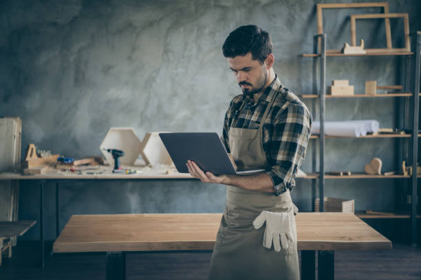 istock Photo of handsome guy holding notebook website administrator reading new orders checking email box wooden business industry woodwork shop garage indoors 1207229100