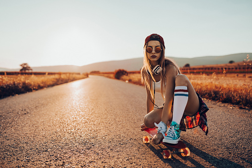 Street styled casual dressed teenage girl with headphones driving skateboard on the road in non-urban scene