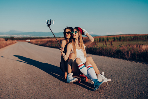 Two playful teenage girls using selfie stick and photographing with smart phone while driving skateboard on a sunny summer day on the road in non-urban scene