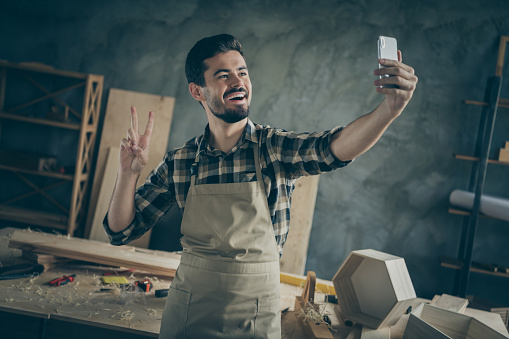 Positive success hardwood furniture restoration entrepreneur workman, make selfie on smartphone show v-sign enjoy his workplace in home house garage