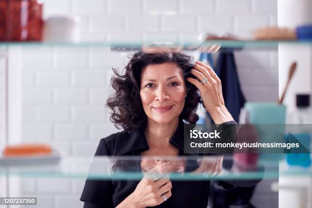 View Through Bathroom Cabinet Of Mature Businesswoman Getting Ready For Work Checking Hair Stock Photo - Download Image Now