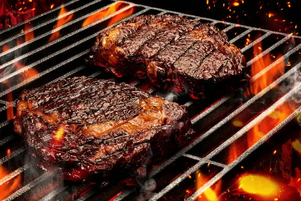 Photo of Process of preparing two pork or beef steaks. Meat roasted on metal portable barbecue BBQ grill with bright flaming fire and ember charcoal. Close up