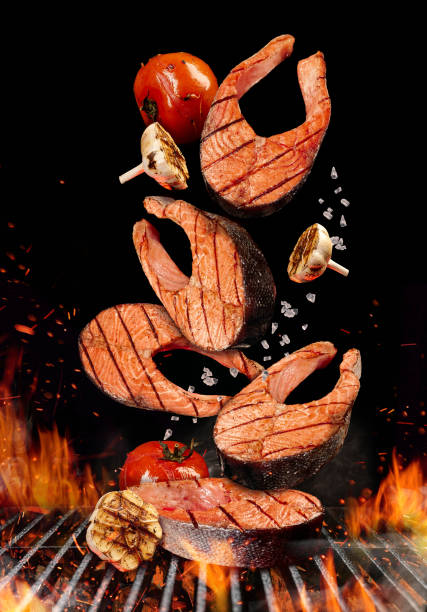 istock Grilled salmon steaks with tomatoes, garlic and salt are falling down on black background. Barbecue bbq grill, fire, charcoal and smoke. Close up 1207250339