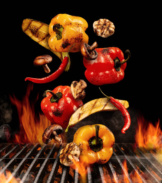 Grilled bell peppers, zucchini, champignons, chilli, halves of garlic are falling down on black background. Bbq grill, flaming fire charcoal. Close up
