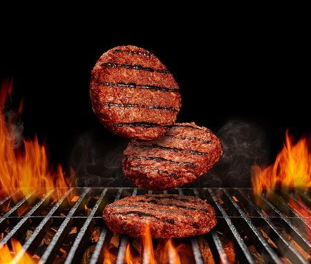 Falling down beef cutlets for burgers. Process of preparing. Meat roasted on metal barbecue BBQ grill with bright flaming fire and ember charcoal on black background. Cookout concept. Close up