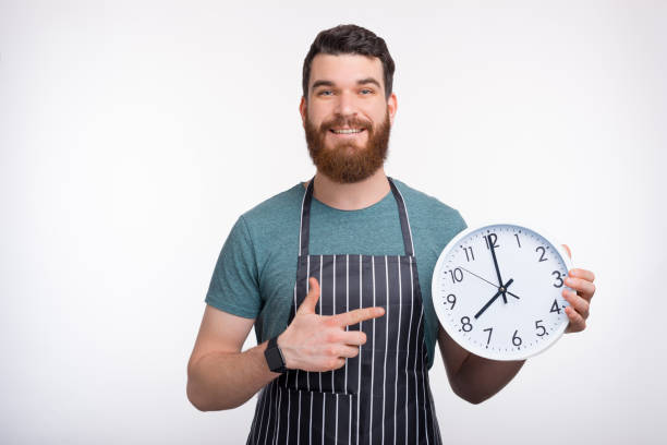 istock Smiling bearded man wearing apron is holding a white clock while smiling at the camera on white background. 1207251772
