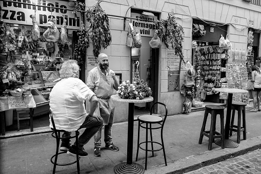Rome, Italy, May 20 - Rome in Black & White series. A tourist sitting outside a gourmet food store near Piazza Navona, in the historic center of Rome, with a great exposition of prosciutto, salami and Italians cheese's.