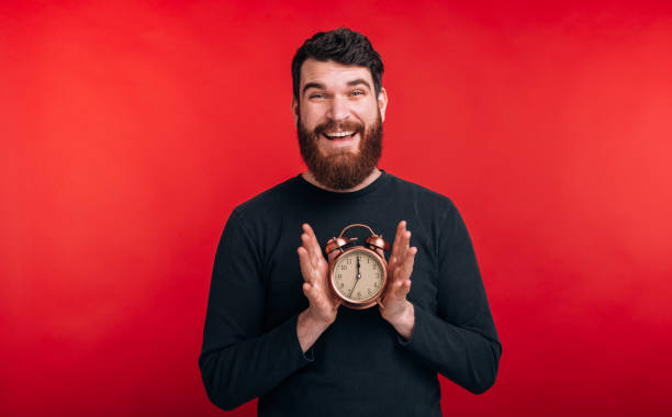 istock It's time. Happy bearded man is holding an alarm clock on red background. 1207252838