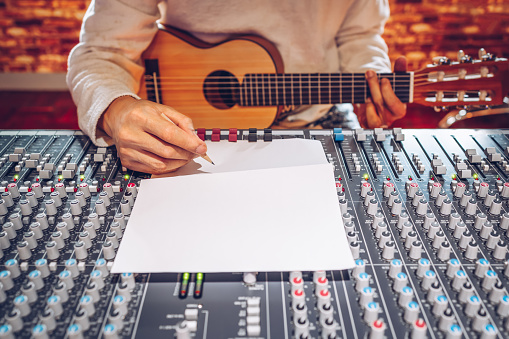 male songwriter playing acoustic guitar and writing a song on audio mixing board in sound studio