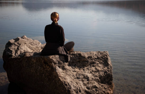 Young woman relaxing by the lake, sitting on a rock in a morning light, watching the beautiful view
