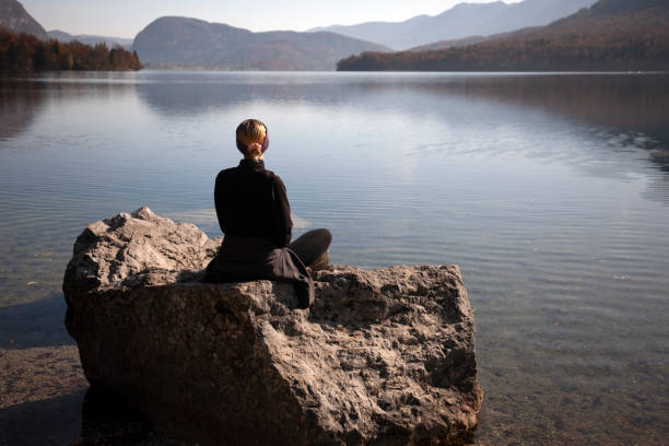 Young woman relaxing by the lake, sitting on a rock in a morning light, watching the beautiful view