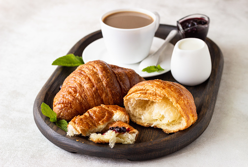 Breakfast concept: two fresh french croissant with a cup of coffee and jam on a dark wooden tray.