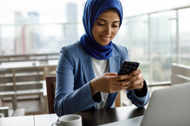 sonriente joven musulmana usando teléfono móvil y computadora portátil en un café - women islam middle eastern ethnicity arabic style fotografías e imágenes de stock