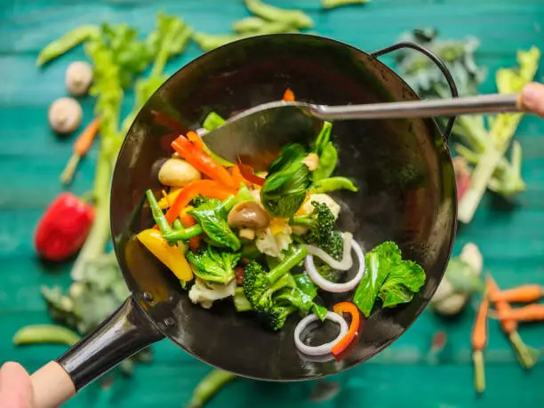 Photo of Stir frying and sauteing a variety of fresh colorful market vegetables in a hot steaming wok with vegetables on on a turquoise colored wood table background below the wok.