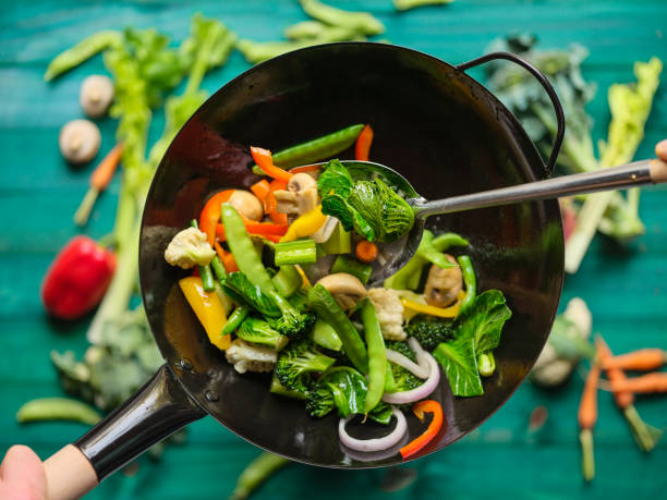 remuer la friture et faire sauter une variété de légumes frais colorés du marché dans un wok chaud à la vapeur avec des légumes sur un fond de table en bois de couleur turquoise sous le wok. - poêle verte photos et images de collection