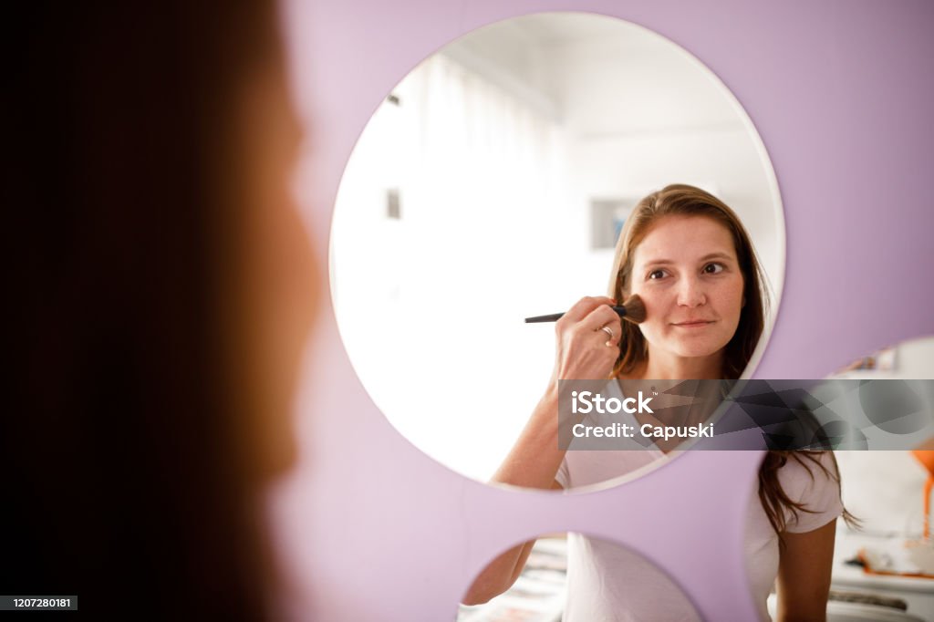 Young woman wearing blush looking in the mirror at bedroom Woman wearing on makeup in front of the mirror at bedroom Mirror - Object Stock Photo