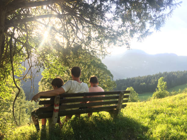 idylliczny widok z tyłu ojca i dwóch synów siedzących na ławce w górach podczas zachodu słońca po wędrówce i odpoczynku. togetherness, bonding, przygoda, zdrowy styl życia, miłość. - mountain mountain climbing climbing snow zdjęcia i obrazy z banku zdjęć