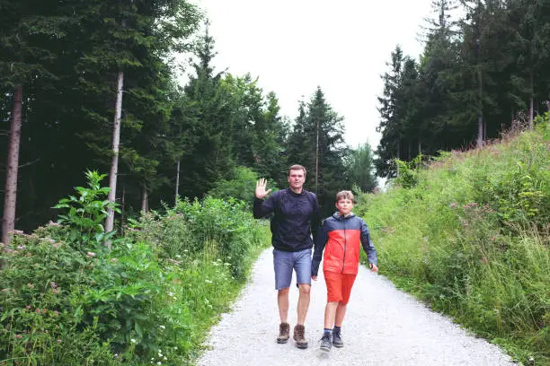 July 27, 2017 - Saalachtal, Austria: dad and son in rain jackets and shorts walking together on a country road among fir trees and tall grass in the forest.
