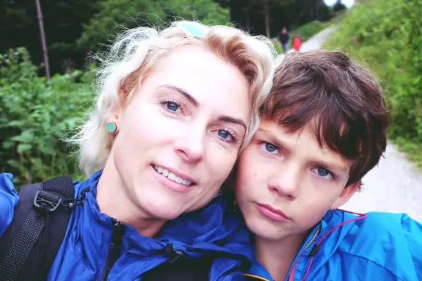 July 27, 2017 - Saalachtal, Austria: selfie of a beautiful blond mother and her boy leaning head next to head, looking at camera while in the forest hiking.