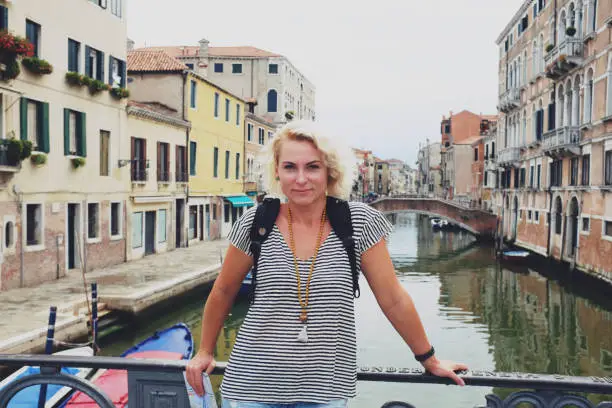 July 30, 2017 - Venice, Italy: beautiful woman in short sleeved shirt, blond hair and necklace standing on a bridge in Venice near canal and empty street, holding map, smiling at camera