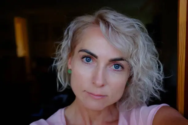 July 20, 2017 - Austria: head and face of a serious pretty blond woman with dark eyebrows and blue eyes looking up curiously from a doorway in the shade in summer.