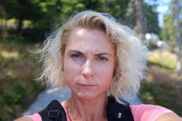 Close up of a head and face of a pretty blond woman with short curly hair making a selfie on hiking trip while being serious, tired and hot stock photo
