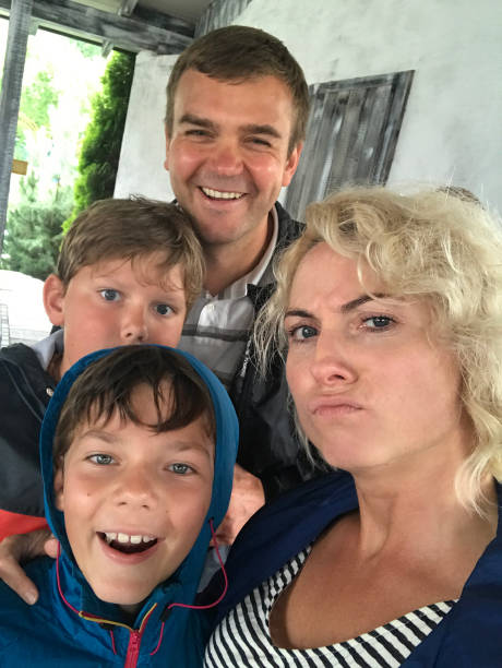 Family selfie - handsome happy father, blond beautiful mother, excited sons posing for fun and cheeky selfie while hiding from rain stock photo