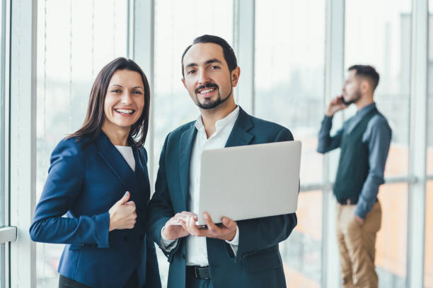 Successful man and woman making business plan, comparing annual data in laptop, while their coleague is on phone on blurred background.