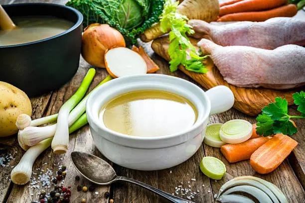Front view of a white bowl filled with a healthy chicken bouillon shot on rustic kitchen table. Ingredients for preparing chicken broth like raw chicken, carrots, kale, potatoes, celery and onions are all around the bowl. High resolution 42Mp studio digital capture taken with SONY A7rII and Zeiss Batis 40mm F2.0 CF lens