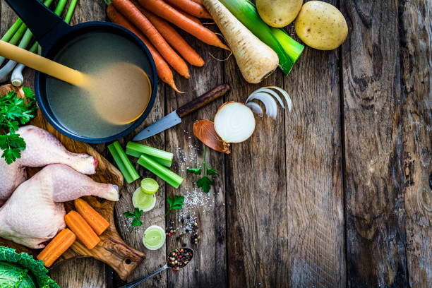 High angle view of a cooking pan filled with a healthy chicken bouillon shot on rustic kitchen table. Ingredients for preparing chicken broth like raw chicken, carrots, kale, potatoes, celery and onions are all around the pan. The composition is at the left of an horizontal frame leaving useful copy space for text and/or logo at the right. High resolution 42Mp studio digital capture taken with SONY A7rII and Zeiss Batis 40mm F2.0 CF lens