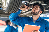 Vehicle service maintenance handsome man checking under car condition in garage. Automotive mechanic pointing flash light on wheel following maintenance checklist document. Car repair service concept
