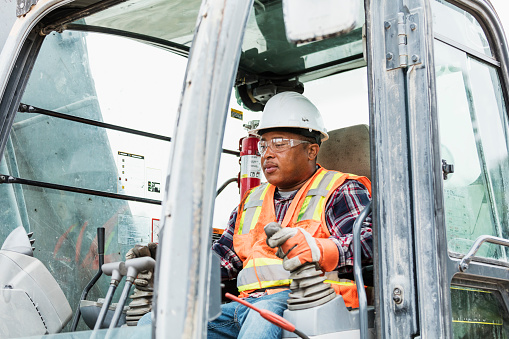 A mature African-American man working at a construction site, driving an earth mover. He is wearing a hardhat, reflective vest, safety goggles and work gloves.