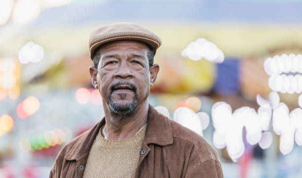 Senior African-American man standing outdoors A senior African-American man in his 60s standing outdoors, looking at the camera. Out of focus in the background are the lights and colorful awnings of a traveling carnival. 65 69 years stock pictures, royalty-free photos & images