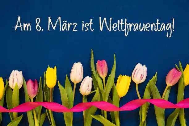 German Text Weltfrauentag Means International Womens Day. White And Pink Tulip Spring Flowers With Ribbon. Blue Wooden Background