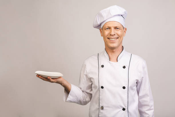 istock Portrait of senior chef holding empty dish isolated on white background. 1207299770